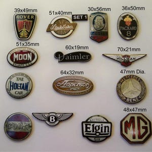 May include: A collection of vintage car badges, including Rover, Studebaker, Lafayette, Packard, Moon, Daimler, Holtan, Apperson, Hillman, Mercedes-Benz, Cord, Bentley, Elgin, and MG. The badges are made of metal and feature a variety of colours and designs.