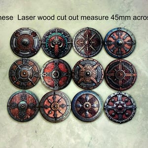 May include: Twelve laser-cut wooden shields with intricate designs. Each shield is 45mm across and features a different pattern in shades of brown, red, and gold.