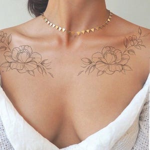 May include: A close-up of a person's chest and shoulders, featuring two large floral temporary tattoos. The tattoos are detailed line drawings of peonies with leaves. A gold triangle pendant necklace is also visible.