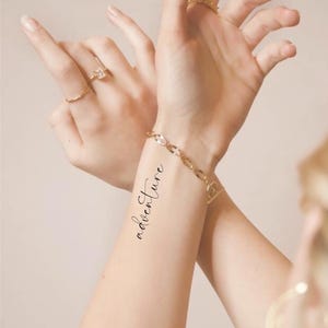 May include: A person's wrist with a temporary tattoo that says "adventure" in cursive script. The person is wearing a gold chain bracelet.