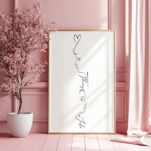 Può includere: Una stampa artistica incorniciata con un disegno a tratto minimalista di due profili e un cuore. L'opera d'arte è in nero su sfondo bianco, in una cornice dorata. I nomi "Tommy" e "Julie" sono scritti in corsivo. La stampa è esposta in una stanza rosa.