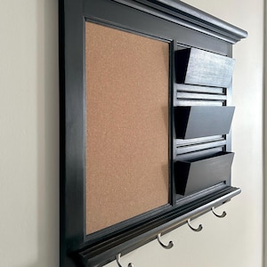 May include: Black wall-mounted organiser with a corkboard, three mail slots, and three hooks.