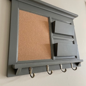 May include: A grey wooden wall organiser with a corkboard, three mail slots, and four hooks. The organiser has a decorative trim around the top and a shelf underneath the hooks.