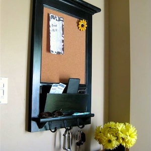 May include: A black framed corkboard with a small note that says "Bread - Milk" and a yellow flower magnet. Below the corkboard is a black shelf with a key rack and sunglasses.