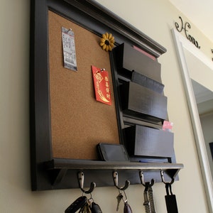 May include: A black wooden wall organiser with a corkboard, mail slots, and hooks. The corkboard has a ticket, a red envelope with Chinese characters, and a yellow flower. The mail slots have envelopes and cards. The hooks have keys and a watch.