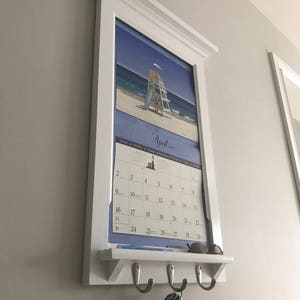 May include: White wooden wall organiser with a shelf and hooks. A calendar with a beach scene is displayed on the organiser. The calendar is for April 2017. A set of keys is hanging on the hooks.