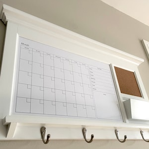 May include: White wall-mounted organizer with a calendar, corkboard, and a small shelf. The calendar has the word "Month" printed at the top and has spaces for each day of the week. The organizer also has three hooks underneath.