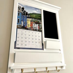 May include: White wall-mounted organizer with a calendar, a chalkboard, a shelf, and three hooks. The calendar features a photo of colorful houses. The chalkboard is blank. The shelf has a white surface. The hooks are gold.