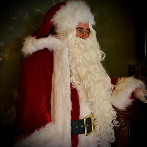 May include: A person dressed as Santa Claus, wearing a red velvet suit with white faux fur trim. The outfit includes a matching hat, a long white beard, and a black belt with a gold buckle. The person is also wearing round glasses.