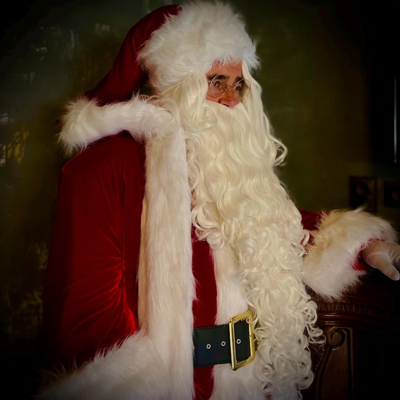 May include: A person dressed as Santa Claus, wearing a red velvet suit with white faux fur trim. The outfit includes a matching hat, a long white beard, and a black belt with a gold buckle. The person is also wearing round glasses.