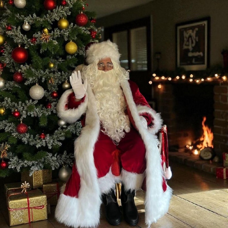 May include: A person dressed as Santa Claus, wearing a red velvet suit with white faux fur trim, waving. A decorated Christmas tree with ornaments and wrapped gifts are in the background. A fireplace with a lit fire is also visible.