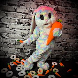 Könnte beinhalten: Ein farbenfrohes Hasenkostüm mit Regenbogen-Tie-Dye-Muster, das eine große orangefarbene Karotte hält. Der Hase hat große Augen und lange Ohren. Spielzeugkarotten und Donuts sind um den Hasen herum verstreut.