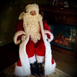 May include: Santa Claus in a red velvet suit with white faux fur trim, a matching hat, and a long white beard. He wears glasses and black boots. The background includes a fireplace and a rug.