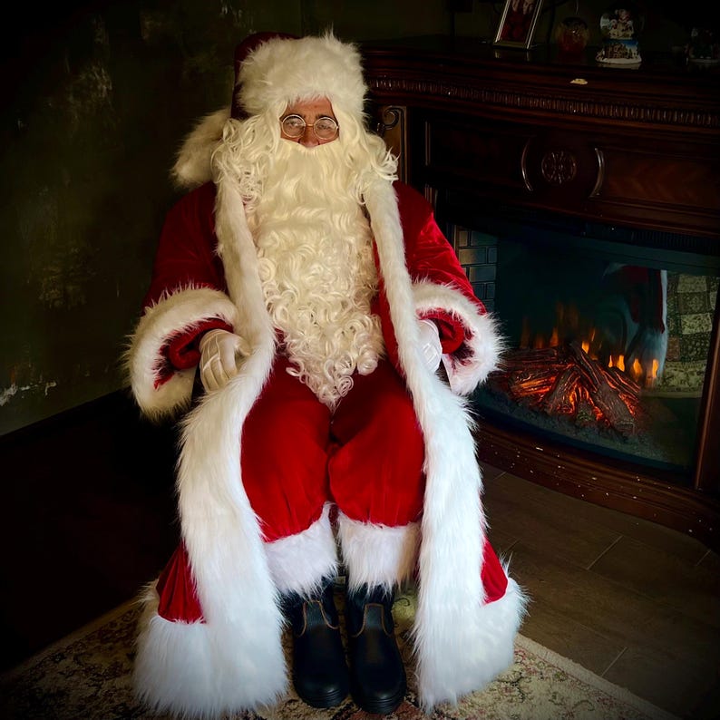 May include: Santa Claus in a red velvet suit with white faux fur trim, a matching hat, and a long white beard. He wears glasses and black boots. The background includes a fireplace and a rug.