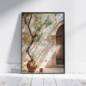 May include: A framed art print showing an olive tree in a terracotta pot, set against a textured, sunlit wall with an arched doorway. The print has a black frame and is displayed on a white wooden floor.