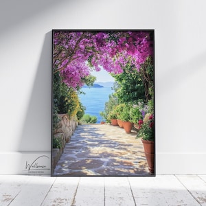 Puede incluir: Impresión enmarcada de un camino de piedra que conduce al mar, enmarcado por vibrantes flores moradas y exuberante vegetación. Macetas de terracota con flores de colores bordean el camino. La impresión se muestra contra una pared blanca y un suelo de madera.