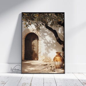 May include: Framed art print featuring a rustic scene with a wooden arched doorway, a large brown jug, and an olive tree. The print has a warm, inviting color palette with shadows and sunlight. The artwork is displayed on a white wall.