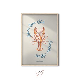 Op de afbeelding: Een ingelijste print met een oranje kreeftillustratie en blauwe cursieve tekst: "Lobster Lover Club Everyday All Day". Het kunstwerk heeft een lichtbeige achtergrond en een houten lijst.