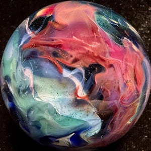 May include: A spherical glass marble featuring a swirling, marbled pattern of vibrant colors including deep red, bright pink, teal, light blue, and white. The surface is smooth and glossy, with tiny internal air bubbles scattered throughout the glass.