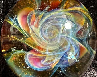May include: A round, glass art piece with a swirling, colorful design. The artwork features a blend of yellow, orange, blue, and pink hues, creating a dynamic and abstract pattern. Small bubbles are scattered throughout the glass.