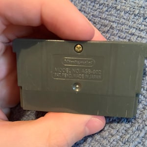 May include: A gray plastic Game Boy Advance game cartridge featuring a central screw and embossed text that reads Nintendo, MODEL NO. AGB-002, PAT. PEND. MADE IN JAPAN. The rectangular cartridge has a notched top edge and a smooth, matte finish.