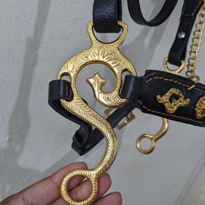 Może przedstawiać: Zbliżenie wędzidła dla konia z ozdobnymi złotymi metalowymi elementami i czarnymi skórzanymi paskami. Wędzidło ma dekoracyjny wzór i łańcuch. Obraz podkreśla kunszt sprzętu jeździeckiego.