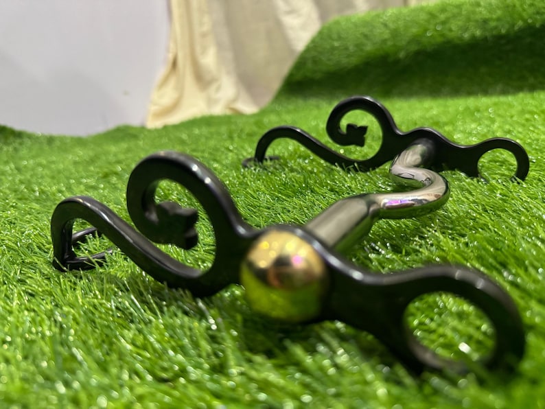 May include: A black and silver horse bit with a decorative scroll design. The bit has a gold-coloured mouthpiece.