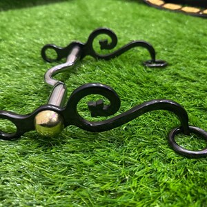 May include: A black metal horse bit with a gold-coloured mouthpiece and decorative swirls. The bit has two rings on either end.