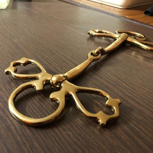 May include: A gold-colored horse bit with a decorative, ornate design. The bit has two rings on one end and a single ring on the other end.