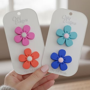 Può includere: Due set di accessori a forma di fiore rosa, arancione, blu e verde acqua, ciascuno montato su una carta bianca. Le carte hanno il testo "Cozy Designs" e "Tiny but lovely".