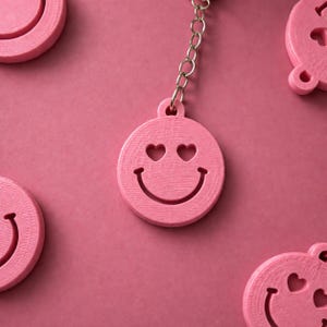 K&ouml;nnte beinhalten: Ein rosa runder Schl&uuml;sselanh&auml;nger mit einem ausgeschnittenen Smiley-Gesicht mit herzf&ouml;rmigen Augen und einem geschwungenen Mund. Der Anh&auml;nger ist an einer silberfarbenen Metallkette befestigt. Mehrere identische Anh&auml;nger liegen um die Mitte.