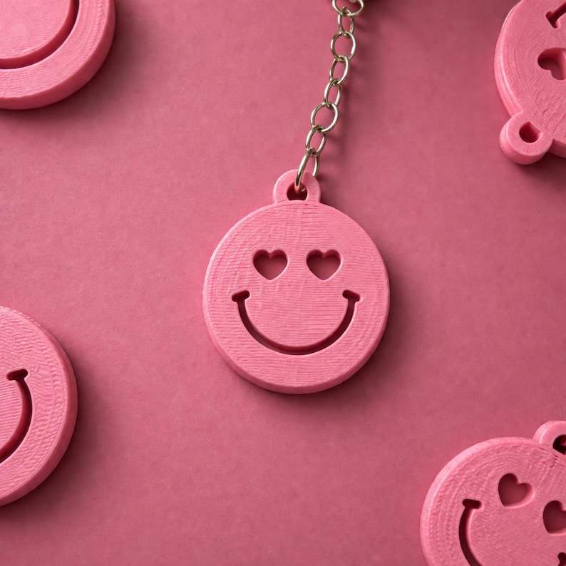 K&ouml;nnte beinhalten: Ein rosa runder Schl&uuml;sselanh&auml;nger mit einem ausgeschnittenen Smiley-Gesicht mit herzf&ouml;rmigen Augen und einem geschwungenen Mund. Der Anh&auml;nger ist an einer silberfarbenen Metallkette befestigt. Mehrere identische Anh&auml;nger liegen um die Mitte.