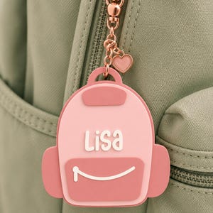 Puede incluir: Un colgante para bolso de silicona con forma de mochila rosa y el nombre Lisa en letras blancas. El accesorio tiene un bolsillo frontal rosa oscuro con una l&iacute;nea curva blanca y est&aacute; unido a una cadena de metal oro rosa con un coraz&oacute;n.