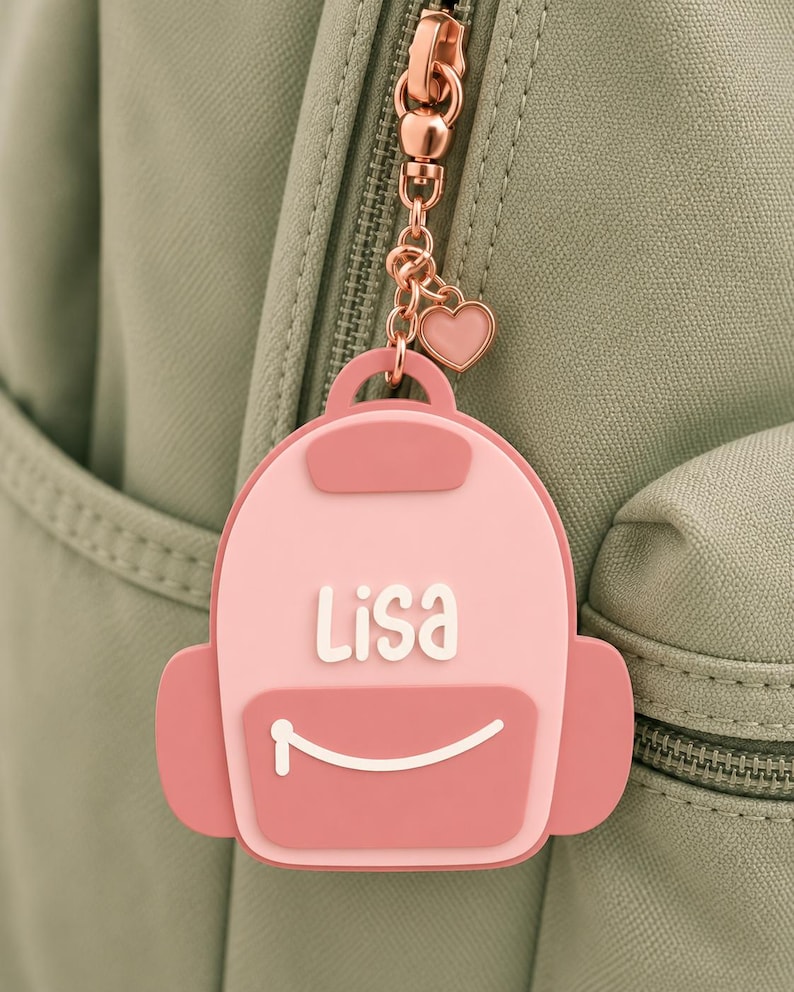 Puede incluir: Un colgante para bolso de silicona con forma de mochila rosa y el nombre Lisa en letras blancas. El accesorio tiene un bolsillo frontal rosa oscuro con una l&iacute;nea curva blanca y est&aacute; unido a una cadena de metal oro rosa con un coraz&oacute;n.