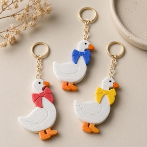 May include: Three white goose keychains with orange beaks and feet, each featuring a different colored bow tie in red, blue, or yellow. The keychains are attached to gold-toned metal rings and chains, showcasing a textured surface on the goose figures.