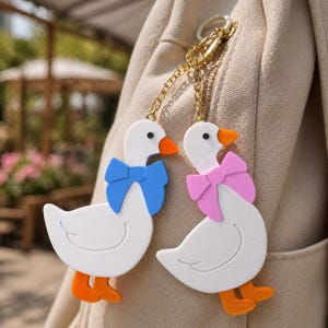 May include: Two white goose keychains with orange beaks and feet, attached to a gold-toned metal chain. One goose wears a blue bow, and the other wears a pink bow. The figures have black eyes and are hanging from a beige fabric surface.
