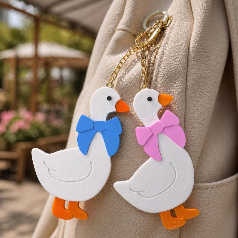 May include: Two white goose keychains with orange beaks and feet, attached to a gold-toned metal chain. One goose wears a blue bow, and the other wears a pink bow. The figures have black eyes and are hanging from a beige fabric surface.