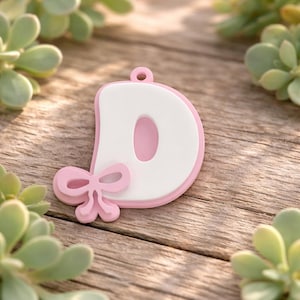 May include: A letter D charm featuring a white center with a pink border and a small pink bow accent attached to the lower left side. The charm includes a small circular loop at the top for hanging, designed with a smooth, flat surface finish.