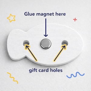 K&ouml;nnte beinhalten: Ein wei&szlig;er, fischf&ouml;rmiger Geschenkkartenhalter mit zwei L&ouml;chern und einem silbernen Magneten. Auf dem Halter steht "Magnet hier ankleben" und "Geschenkkartenl&ouml;cher". Gelbe Pfeile zeigen auf die L&ouml;cher. Dekorative gelbe und blaue Formen umgeben den Halter.