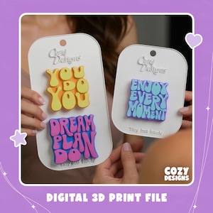 Puede incluir: Dos tarjetas de presentación blancas con letreros de plástico impresos en 3D. La tarjeta izquierda muestra texto amarillo YOU DO YOU y rosa DREAM PLAN DO. La tarjeta derecha muestra texto azul ENJOY EVERY MOMENT. Fuente de estilo ondulado.