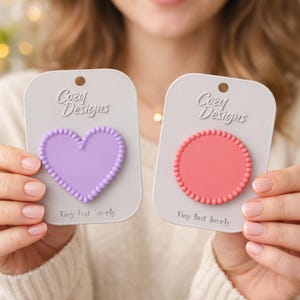 May include: Two decorative phone grips are held up. One is a light purple heart shape, and the other is a coral pink circle. Both have a beaded border and are on a gray card with the text "Cozy Designs" and "Tiny but lovely".