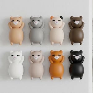 May include: A collection of eight cat-shaped magnets in various colors, including tan, gray, cream, brown, white, and black. Each magnet features a minimalist design with arms raised to the head, and a small pink nose.