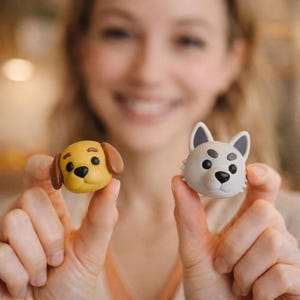 K&ouml;nnte beinhalten: Zwei kleine, hundef&ouml;rmige Figuren. Eine ist gelb mit braunen Ohren, die andere grau mit schwarzen Akzenten. Beide haben schwarze Augen und Nasen, vor einem unscharfen Hintergrund.
