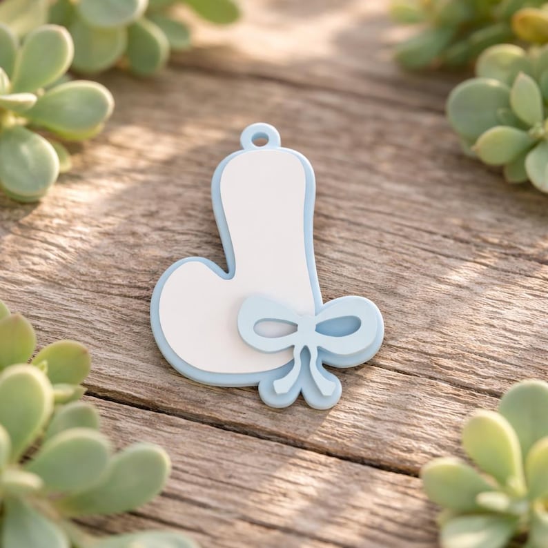 May include: A light blue and white letter J pendant featuring a smooth finish and a decorative bow accent at the base. The letter has a rounded shape with a small loop at the top for hanging, showcasing a layered design with a white center.
