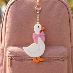 May include: A white goose keychain with an orange beak and feet, wearing a pink bow around its neck. The charm is attached to a gold-toned metal chain featuring a small flower-shaped link, hanging against a dusty rose-colored fabric backpack.