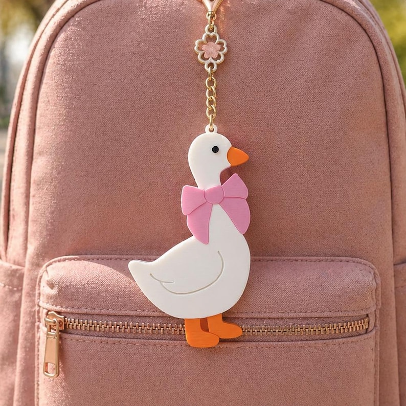 May include: A white goose keychain with an orange beak and feet, wearing a pink bow around its neck. The charm is attached to a gold-toned metal chain featuring a small flower-shaped link, hanging against a dusty rose-colored fabric backpack.