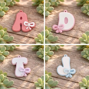 May include: Four individual letter charms featuring the letters A, D, T, and J. Each letter has a rounded, puffy design with a small attached bow detail. The letter A is coral, D is white with a pink border, T is white with a pink border, and J is white with a light blue border.