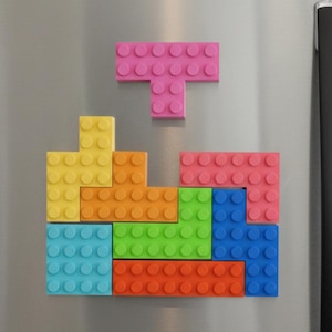 May include: A colorful arrangement of magnetic building block toys in various shapes and colors, including pink, yellow, orange, green, blue, and red. The blocks are arranged on a silver surface, resembling a Tetris game.