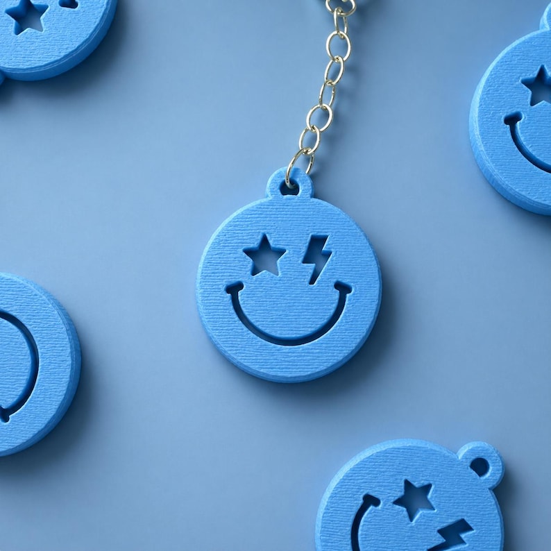 K&ouml;nnte beinhalten: Ein hellblauer, kreisf&ouml;rmiger Schl&uuml;sselanh&auml;nger mit einem ausgeschnittenen Smiley-Gesicht, bei dem ein Auge als Stern und das andere als Blitz geformt ist. Der Anh&auml;nger ist an einer silberfarbenen Metallkette befestigt und hat eine matte Oberfl&auml;che.
