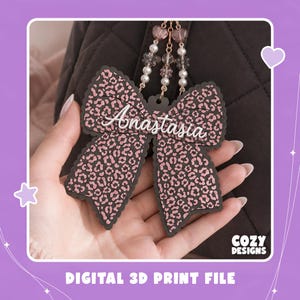 Könnte beinhalten: Ein schleifenförmiger Taschenanhänger mit schwarz-rosa Leopardenmuster und dem Namen Anastasia in weißer Schreibschrift. Der Anhänger ist an einer goldfarbenen Kette mit kleinen weißen Perlen und rosa herzförmigen Akzenten befestigt.